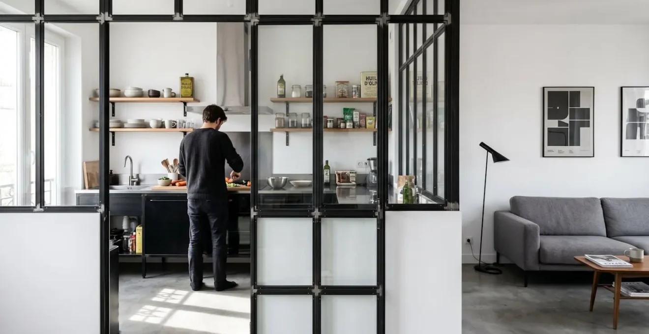 Verrière style atelier à cadre noir séparant une cuisine d'un salon dans un appartement rénové, lumière naturelle traversant les carreaux de verre