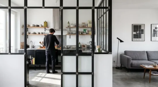 Verrière style atelier à cadre noir séparant une cuisine d'un salon dans un appartement rénové, lumière naturelle traversant les carreaux de verre